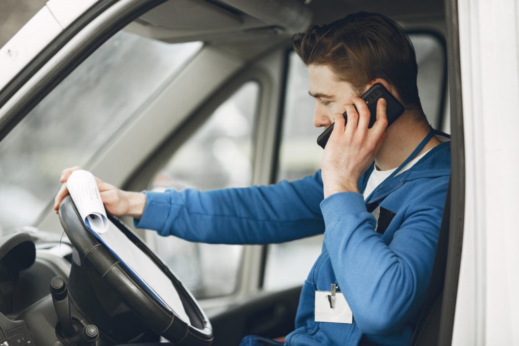 Homem sentado no banco do caminhão, vestido com uniforme de entrega, falando ao celular e segurando prancheta.