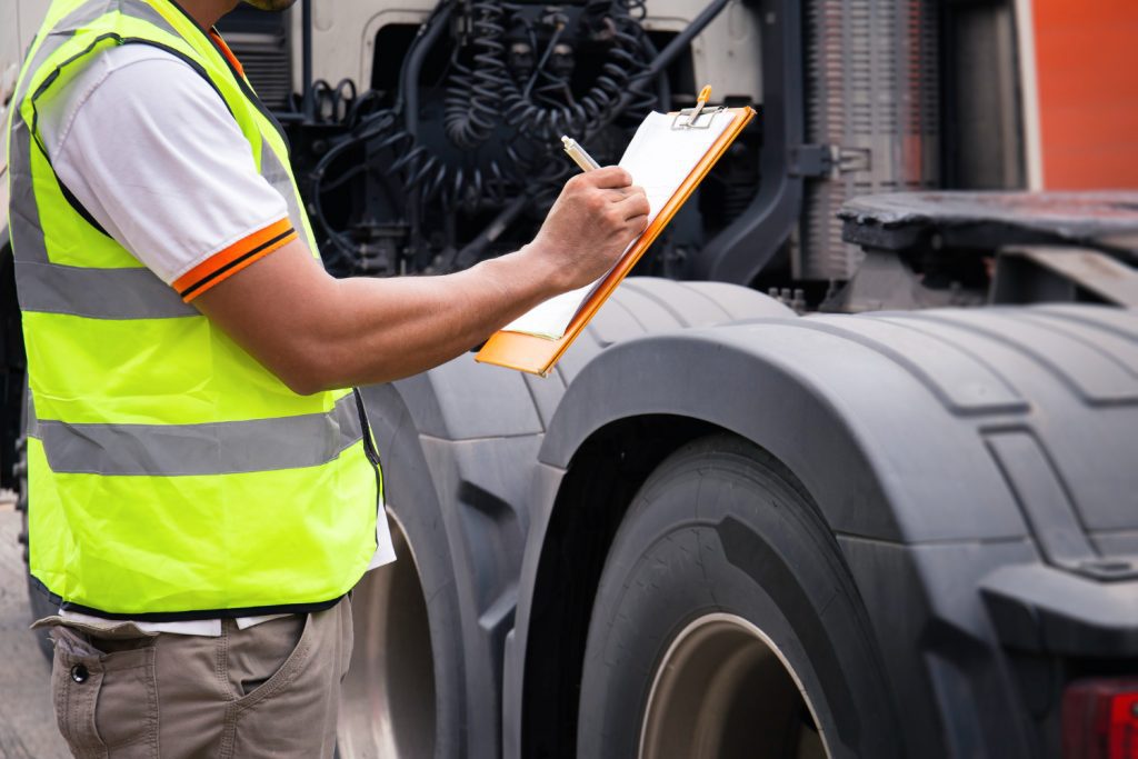 Homem segura prancheta verificando dados sobre um caminhão de transporte de carga.